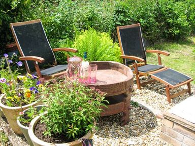 A garden seating area with two wooden lounge chairs a round table with bottles and glasses and several potted plants at Lothlorien Cottage in Cwmgors near Ammanford