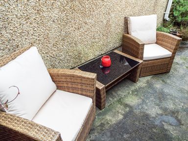 Two wicker chairs with white cushions and a small glass-top table with a red candle on a concrete patio at Lothlorien Cottage in Cwmgors near Ammanford