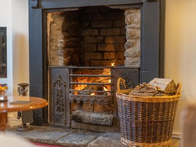 A lit fireplace with a metal grate and a wicker basket filled with firewood beside it at The Coach House in Gilwern near Crickhowell and Abergavenny