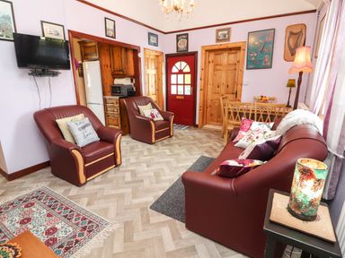 A living room with brown leather chairs and sofa a red door wooden dining table and chairs a wall-mounted television and kitchen visible at Brendan's Cottage in Valentia Island County Kerry
