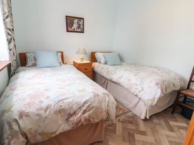 A bedroom with two single beds a wooden nightstand with a lamp a chair and a painting on the wall at Brendans Cottage Valentia Island County Kerry