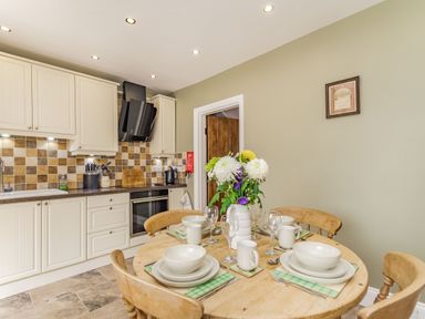 A kitchen with a round wooden table set for four and a kitchenette with cream cabinets and a tiled backsplash at Fisher Cottage in Hornsea