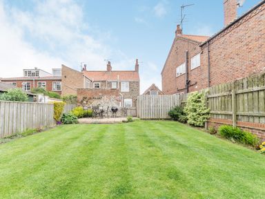 A backyard with a grass lawn surrounded by wooden fences and a patio with a table, chairs, and a barbecue at Fisher Cottage in Hornsea