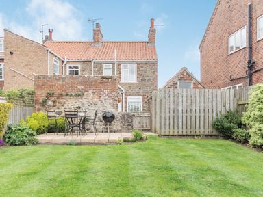 A backyard with a grass lawn a paved seating area with chairs and a table a charcoal grill a brick and stone wall and wooden fences at Fisher Cottage in Hornsea