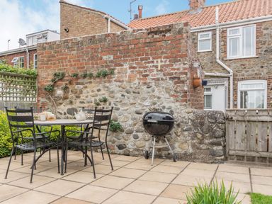 A patio with a metal table and chairs and a charcoal grill against a brick and stone wall at Fisher Cottage in Hornsea