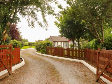 A gravel driveway with open wooden gates leading to a house surrounded by trees and hedges at The Lodge in Churchtown near Beaufort County Kerry