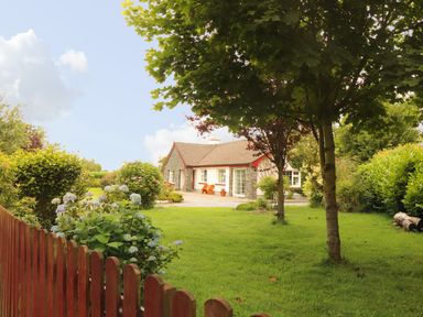 A stone and white cottage with a garden and trees behind a wooden fence at The Lodge Churchtown near Beaufort County Kerry