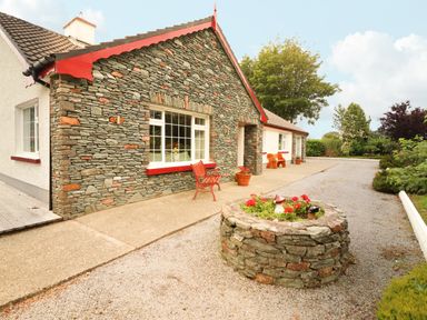 A stone and white house with red trim a red bench and a circular stone flower bed on a gravel driveway at The Lodge Churchtown near Beaufort County Kerry