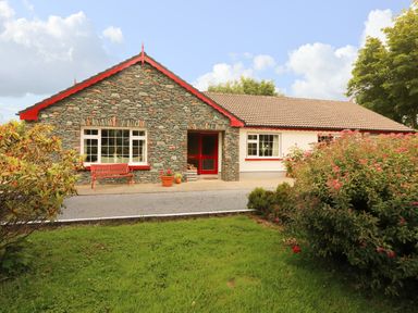 A stone and white house with red trim surrounded by green grass and bushes at The Lodge in Churchtown near Beaufort County Kerry