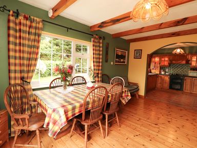 A dining room with wooden chairs and a table covered with a plaid tablecloth next to a window with matching curtains and a visible kitchen through an archway at The Lodge in Churchtown near Beaufort County Kerry