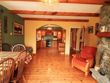 A dining area with wooden chairs and a table with a checkered tablecloth next to a lounge chair and a stone fireplace with a kitchen visible through an archway at The Lodge in Churchtown near Beaufort County Kerry