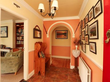 A hallway with framed pictures on the wall a wooden bench a mirror a vase with flowers and an arched doorway showing a living room with bookshelves at The Lodge in Churchtown near Beaufort County Kerry