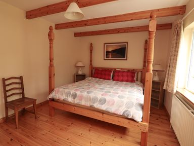 A bedroom with a wooden four-poster bed a wooden chair two bedside tables with lamps a window and a framed picture on the wall at The Lodge in Churchtown near Beaufort County Kerry