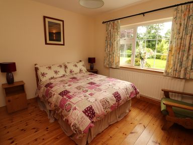 A bedroom with a double bed beside two side tables with lamps and a window showing a garden outside at The Lodge in Churchtown near Beaufort County Kerry