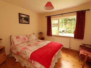 A bedroom with a double bed with patterned bedding and red throw a window with red curtains wooden floor a framed picture on the wall at The Lodge in Churchtown near Beaufort County Kerry