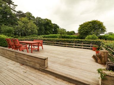 An outdoor wooden deck with red table and chairs surrounded by hedges and trees at The Lodge in Churchtown near Beaufort County Kerry