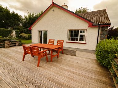 An outdoor wooden deck with a wooden table and four chairs beside a white house with a window and sliding glass door at The Lodge Churchtown near Beaufort County Kerry
