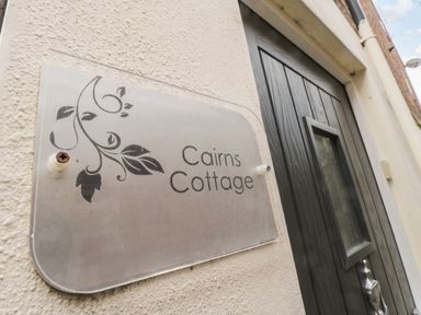 A sign with leaves and the text Cairns Cottage on a wall near a dark wooden door at Cairns Cottage in Whitby