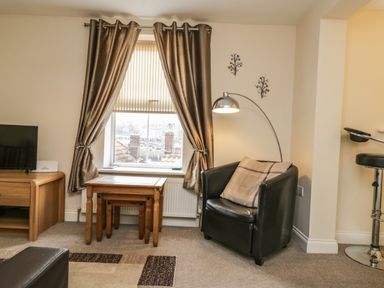 A living room with a black armchair pillow wooden tables window with curtains a floor lamp wooden TV stand and a bar stool at Cairns Cottage in Whitby