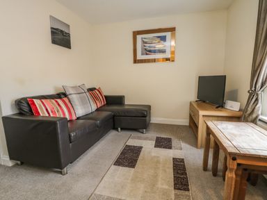 A living room with a black leather sofa with cushions a wooden table a television on a wooden stand and two pictures on the walls at Cairns Cottage in Whitby