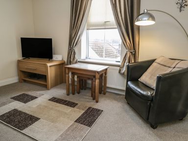 A living room with a television on a wooden stand wooden nesting tables a black armchair with a cushion and a floor lamp at Cairns Cottage in Whitby