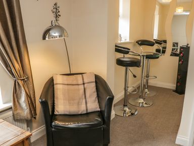 A corner seating area with a black leather armchair with a cushion a metallic floor lamp bar stools and a tall speaker at Cairns Cottage in Whitby