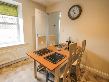A dining area with a wooden table set with four chairs placemats wine glasses and a bottle a wall clock and a window with a green blind at Cairns Cottage in Whitby
