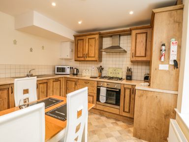 A kitchen with wooden cabinets a gas stove a microwave and a dining table with white chairs at Cairns Cottage in Whitby