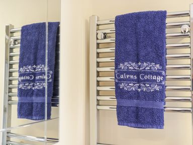 A blue towel with Cairns Cottage embroidered on a chrome towel rack with a mirror reflecting the towel at Cairns Cottage in Whitby