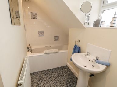 A bathroom with a bathtub under a sloped ceiling a pedestal sink a radiator and a window at Cairns Cottage in Whitby