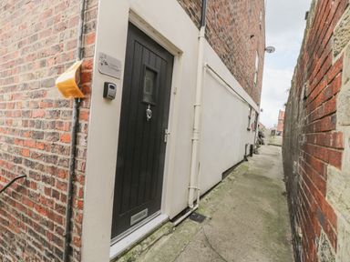A black door with a mail slot and door knocker in a narrow alleyway between brick walls at Cairns Cottage in Whitby