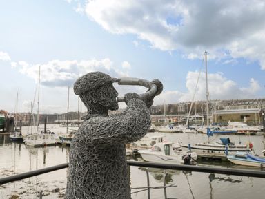 A wire sculpture of a person playing a trumpet by a marina with boats at Cairns Cottage in Whitby