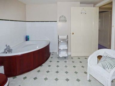 A bathroom with a corner bathtub wooden panel and white wicker chair with cushion at The White Cottage in Bridlington