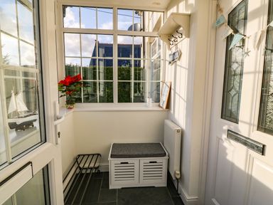 A small enclosed porch with a white storage bench a window shelf with a flower pot and decorations at Ty'r Ysgol in Morfa Bychan