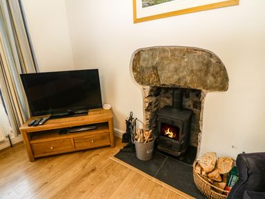 A corner of a room with a wooden TV stand holding a flat screen TV and remote controls next to a stone fireplace with a wood burning stove and firewood at Ty'r Ysgol in Morfa Bychan
