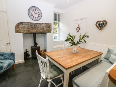 A dining area with a wooden table surrounded by white chairs and bench seating a small stove under a large stone mantle and a clock above at Ty'r Ysgol in Morfa Bychan