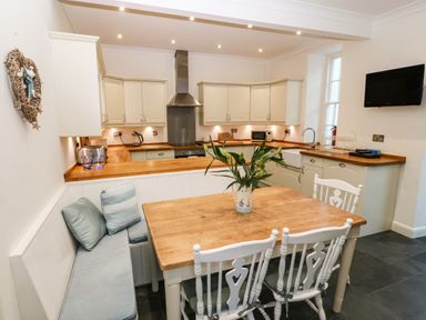 A kitchen with white cabinets wooden countertops a dining table with white chairs and a bench with cushions at Ty'r Ysgol Morfa Bychan