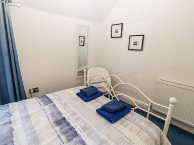 A bedroom with a bed with blue and white bedding two folded blue towels on the bed a white chair a wall mirror two framed pictures and a radiator at Ty'r Ysgol in Morfa Bychan