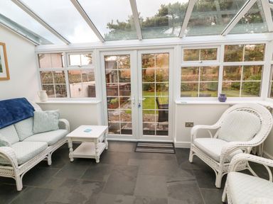 A conservatory with wicker chairs a small table and glass doors opening to a garden at Ty'r Ysgol in Morfa Bychan