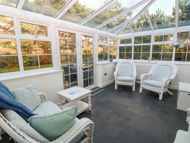 A conservatory with white wicker furniture a small white table and large windows showing trees at Ty'r Ysgol in Morfa Bychan