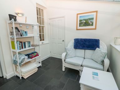 A small seating area with a white wicker loveseat with cushions and a blanket a white shelf filled with books blankets and cushions and a white door at Ty'r Ysgol in Morfa Bychan