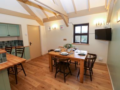 A dining area with a wooden table set for four people and wooden chairs in a room with exposed ceiling beams a window and a mounted television at Foxglove in Newport Pembrokeshire
