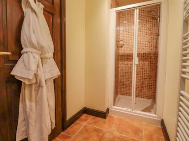 A bathroom with a shower enclosure with brown tiled walls two white bathrobes hanging on a wooden door and a white radiator on the wall at Dairy Barn in Pershore