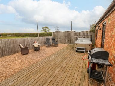 An outdoor patio with wooden decking containing a barbecue grill and hot tub and gravel area with chairs and table at Dairy Barn in Pershore