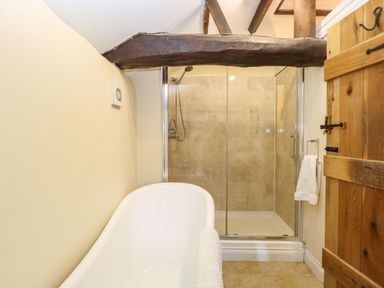A bathroom with a white bathtub exposed wooden ceiling beams a glass shower enclosure and a wooden door at The Barn Saham Hills near Swaffham