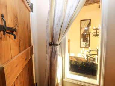 A wooden door next to a tied back curtain and a mirror reflecting a chandelier and furniture at The Barn Saham Hills near Swaffham