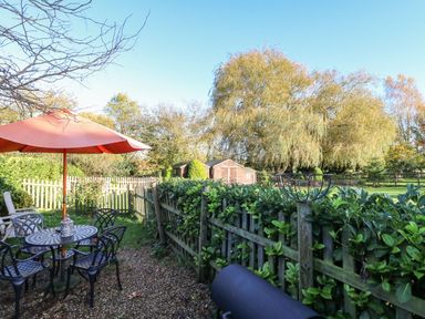 A garden with a table and chairs under an umbrella and a wooden fence with trees in the background at The Barn Saham Hills near Swaffham