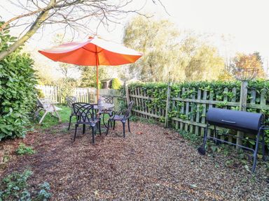 A garden area with a round table and four chairs under an orange umbrella near a wooden fence and barbecue grill at The Barn Saham Hills near Swaffham