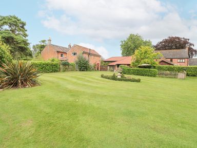 A large lawn with trimmed hedges and ornamental bushes in front of brick houses with tiled roofs at The Barn Saham Hills near Swaffham