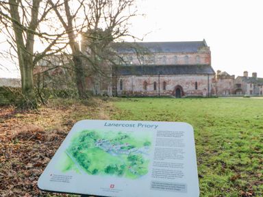 An outdoor scene with a historical priory building and an information sign about Lanercost Priory in Banks near Brampton